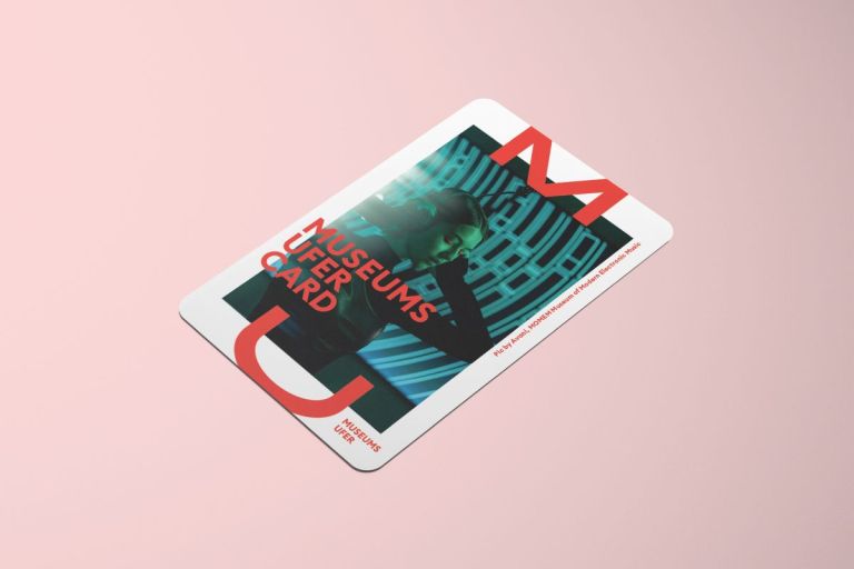 Die MuseumsuferCard mit einem Foto, auf dem eine junge Frau einen Kopfh&ouml;rer tr&auml;gt und sich mit den H&auml;nden an die Ohren fasst in dunklem Raum, auf rosa Hintergrund